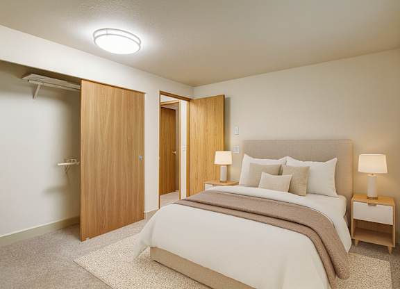 A calm bedroom here at Island Park featuring a bed with neutral bedding and layered pillows, a soft area rug over plush carpeting, and matching bedside tables with modern lamps. The space includes a sliding closet with wood doors, light-colored walls, and a wide window with blinds that brings in natural light.