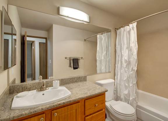 A clean bathroom here at Island Park featuring a wide vanity with a stone-look countertop, integrated sink, and wood cabinetry beneath a large wall-mounted mirror and modern light fixture. The space includes a tub with a white shower curtain, a toilet, neutral wall tones, and towel hardware creating a simple, functional layout.