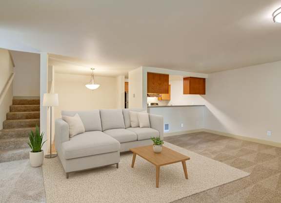 A spacious living room here at Island Park featuring neutral carpeting, a light sectional sofa with accent pillows, and a simple wooden coffee table on an area rug. The open layout includes a staircase with carpeted steps, a floor lamp, and sightlines into the adjacent kitchen and dining area with warm wood cabinetry.