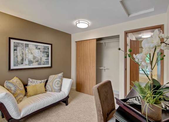 A cozy den here at Island Park featuring soft carpet flooring, a white cushioned futon with patterned accent pillows, and a neutral accent wall with framed artwork. The space includes a built-in closet with sliding wood doors, a compact desk with a chair, overhead lighting, and an open doorway leading to the adjacent living area.