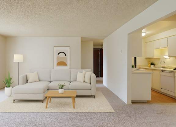 A staged living room here at Lakeside featuring a light neutral sectional sofa with accent pillows, a wooden coffee table on a textured area rug, and a floor lamp beside a potted plant, with an abstract wall art piece above the sofa and an open view into a bright kitchen with white cabinetry and countertops.