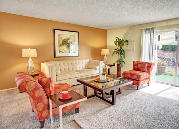 A welcoming living room here at Lakeside featuring plush carpet, a neutral sofa paired with patterned accent chairs, a glass-top coffee table, and warm table lamps, with framed wall art, a tall indoor plant, and sliding glass doors that bring in natural light and connect the space to a private outdoor patio area.