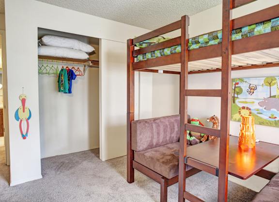 A cheerful bedroom here at Lakeside featuring soft carpet, a wooden loft bed with a built-in desk and seating area below, playful bedding and décor, and an open closet with shelving and hanging space, creating a functional layout that maximizes floor space while offering a cozy and organized environment.