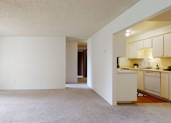 A spacious living room here at Lakeside featuring neutral walls, plush carpet flooring, and an open layout that flows into a bright kitchen with ample cabinetry and countertop space, while a sliding glass door brings in natural light and provides access to an outdoor area, creating a flexible and inviting living space.