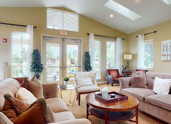A welcoming resident lounge here at Lakeside featuring plush sofas with accent pillows, a round wooden coffee table, upholstered chairs, and soft carpeting, with tall windows, skylights, and glass doors that bring in natural light while offering views of the surrounding community and outdoor seating area.