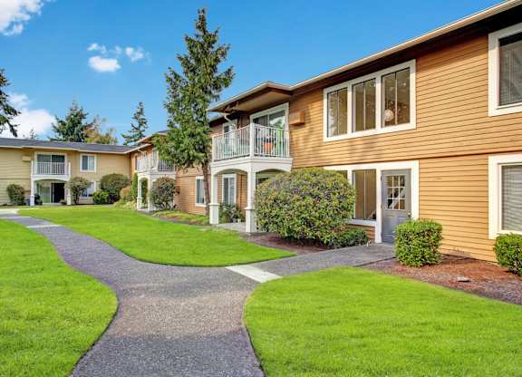 A landscaped courtyard here at Lakeside showcasing two-story apartment buildings with warm-toned siding, private balconies and patios, neatly trimmed shrubs, mature trees, and lush green lawns, with a winding paved walkway leading through the community beneath a bright blue sky.