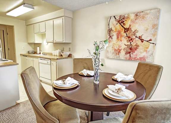 A cozy dining area here at Lakeside showcasing a round dining table set for four with upholstered chairs, neutral carpeting, and a decorative centerpiece, complemented by soft wall art and an adjacent kitchen with light cabinetry, ample counter space, and overhead lighting that creates a warm, inviting flow between spaces.