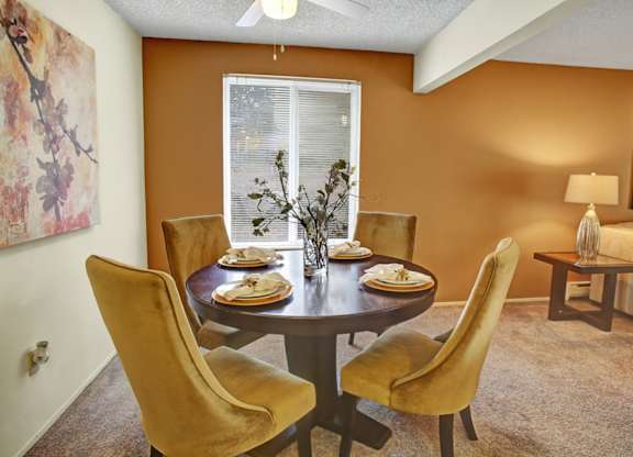A warm dining area here at Lakeside featuring a round wooden dining table set for four with upholstered chairs, soft neutral carpeting, and an accent wall in a rich earth tone, complemented by a ceiling fan, natural light from a nearby window, and a cozy seating area with side table and lamp visible in the background.