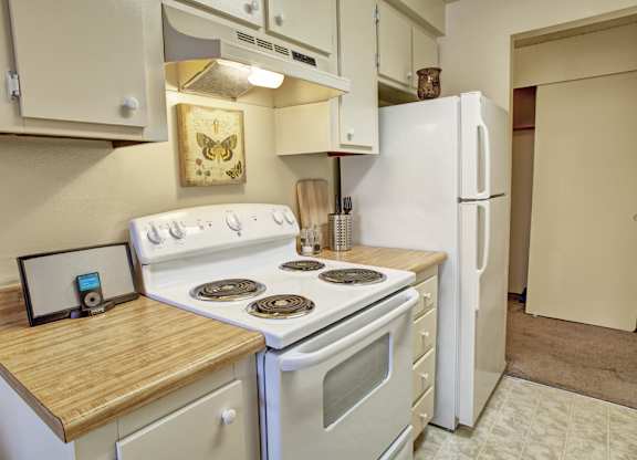 A cozy kitchen here at Lakeside featuring light-toned cabinetry, wood-style countertops, and a clean cooking area with a range, refrigerator, and overhead storage, accented by simple wall art, small countertop décor, and an open closet doorway that adds depth and functionality to the space.
