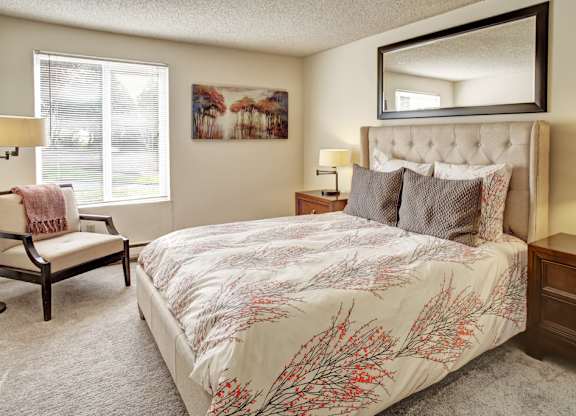 A welcoming bedroom here at Lakeside showcasing a plush bed with patterned bedding and layered pillows, flanked by matching wood nightstands with table lamps, complemented by a cozy accent chair, soft carpet flooring, wall art, and a large window with blinds that brings in natural light.