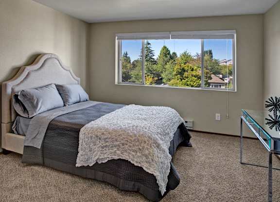 A bright bedroom here at Leilani on Greenwood featuring soft carpeted flooring, neutral walls, and a bed with layered gray and white bedding. A large window with blinds fills the room with natural light and offers leafy neighborhood views, while the open layout provides comfortable space for bedroom furnishings.