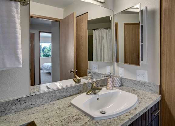 A clean bathroom here at Leilani on Greenwood featuring a light stone countertop with an integrated sink, a wood vanity with storage below, and a wide wall mirror with overhead lighting. Neutral walls, coordinating fixtures, and a nearby doorway create a bright, functional layout connected to the bedroom.