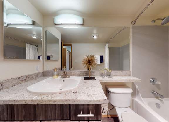 A well-appointed bathroom here at Leilani on Greenwood featuring a wood vanity with a light stone countertop, an integrated sink, and wide wall mirrors with overhead lighting. A toilet sits beside the vanity, while a bathtub-shower combo with a curtain completes the clean, functional layout.