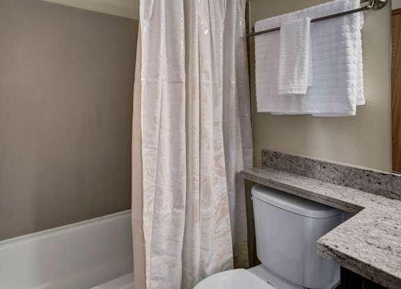 A simple bathroom here at Leilani on Greenwood featuring a bathtub-shower combo with a curtain, a toilet positioned beside a light stone countertop, and neutral wall finishes. A towel bar with folded towels and clean lines create a practical, easy-to-use space within the apartment.