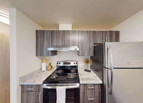 A modern kitchen here at Leilani on Greenwood featuring gray wood-style cabinetry, light stone countertops, and a full cooking area with a range, hood, and refrigerator. Upper and lower cabinets provide storage, while clean finishes, simple décor, and overhead lighting create a functional and contemporary kitchen layout.