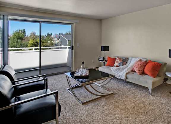 A spacious open living area here at Leilani on Greenwood featuring soft carpeted flooring, neutral walls, and an open layout that connects to the kitchen. A breakfast bar with a light stone countertop overlooks the living space, while overhead lighting and a hallway entry create a clean, functional apartment layout.