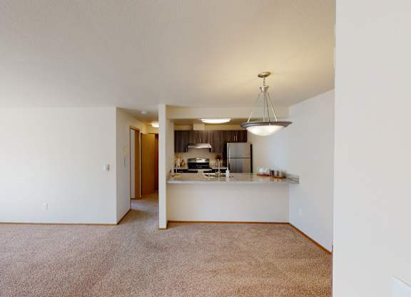 A spacious open living area here at Leilani on Greenwood featuring soft carpeted flooring, neutral walls, and an open layout that connects to the kitchen. A breakfast bar with a light stone countertop overlooks the living space, while overhead lighting and a hallway entry create a clean, functional apartment layout.