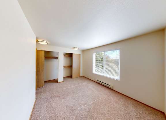 A spacious bedroom here at Leilani on Greenwood featuring soft carpeted flooring, neutral walls, and a large window with blinds that brings in natural light. Two open closets with hanging rods provide ample storage, while overhead lighting and a simple layout create a bright, comfortable sleeping space.