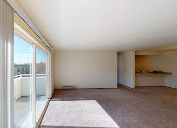 A sunlit open living area here at Leilani on Greenwood featuring soft carpeted flooring, neutral walls, and large sliding glass doors with vertical blinds that open to a private balcony. Natural light fills the space, which flows toward the kitchen area with a breakfast bar, creating an airy and inviting layout.