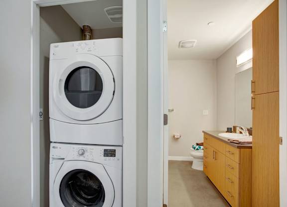 A convenient laundry nook here at Leilani on Greenwood featuring a stacked washer and dryer tucked inside a dedicated closet, positioned just off a bathroom with a long vanity, warm wood cabinetry, a smooth countertop with an integrated sink, and overhead lighting, creating a functional and efficient in-home layout.