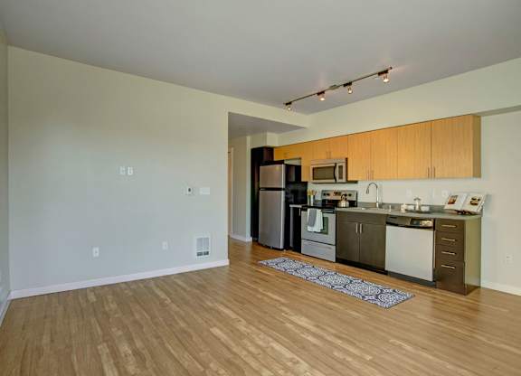 A spacious open living area here at Leilani on Greenwood featuring wood-style flooring, a wide wall of space for flexible furniture placement, and a modern kitchen with warm wood cabinetry, a full appliance suite, sleek countertops, and track lighting that brightens the open-concept layout.