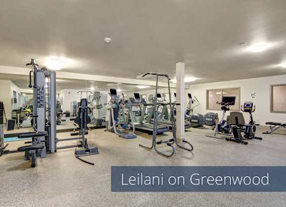A spacious fitness center here at Leilani on Greenwood featuring a variety of cardio machines, strength training equipment, and free weights arranged throughout an open, well-lit room with neutral flooring and ceiling lights, creating a convenient on-site workout space for residents.