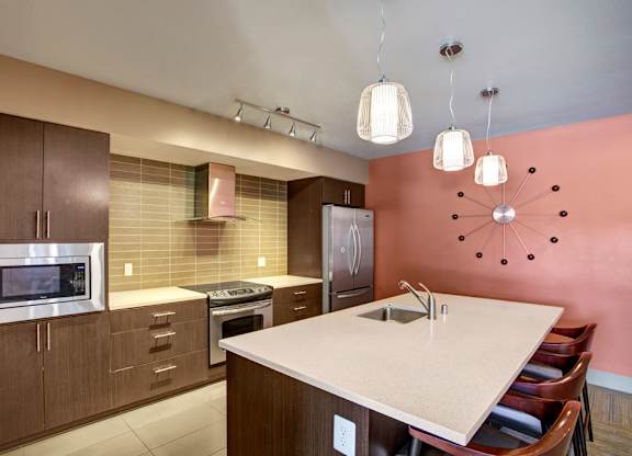 A modern community kitchen here at Leilani on Greenwood featuring an oversized island with bar seating, sleek countertops, contemporary pendant lighting, and a full appliance suite framed by dark wood cabinetry, with a tiled backsplash and bold accent wall that create a stylish space for resident gatherings and events.