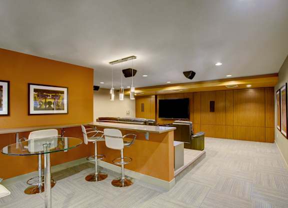 A contemporary media lounge here at Leilani on Greenwood featuring bar-height counter seating with modern stools, a glass-top table, warm accent walls with framed artwork, and a spacious viewing area with comfortable seating and a large mounted television designed for entertaining, gatherings, and movie nights.