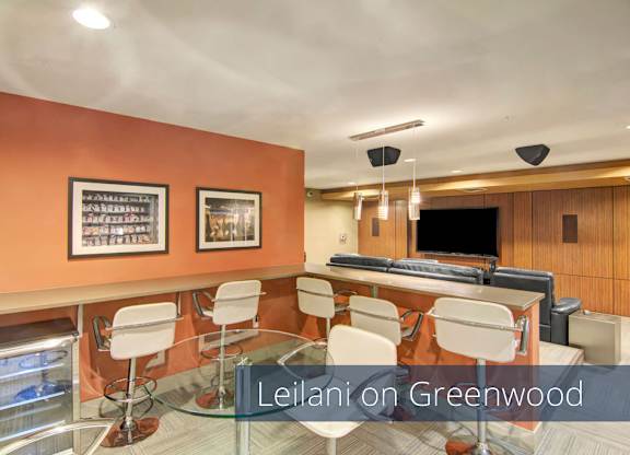 A contemporary media lounge here at Leilani on Greenwood featuring bar-height counter seating with modern stools, a glass-top table, warm accent walls with framed artwork, and a spacious viewing area with comfortable seating and a large mounted television designed for entertaining, gatherings, and movie nights.