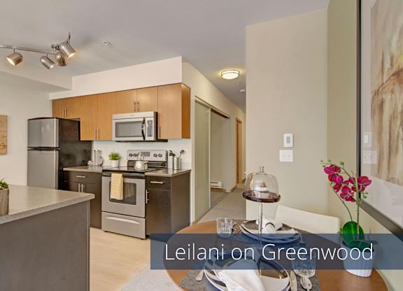 A contemporary kitchen and dining area here at Leilani on Greenwood featuring flat-panel wood cabinetry, dark lower cabinets, smooth countertops, and a stainless steel sink and appliances. A peninsula counter opens to a dining table, while track lighting and light wood-style flooring create a clean, modern layout.