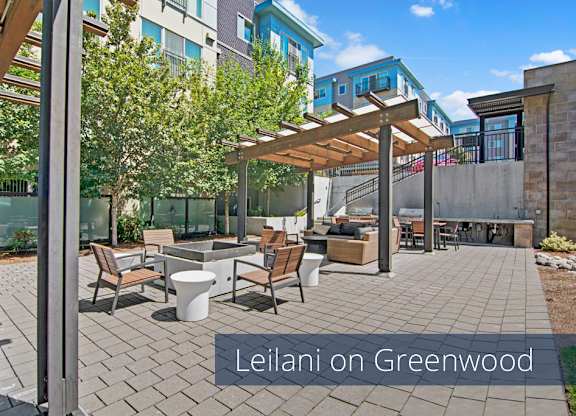 A landscaped outdoor courtyard here at Leilani on Greenwood featuring a paved patio with modern seating, wood pergolas casting shade over lounge areas, cushioned sofas, café-style chairs and tables, built-in fire pit seating, and grilling stations, all surrounded by mature trees, privacy fencing, and contemporary apartment buildings.