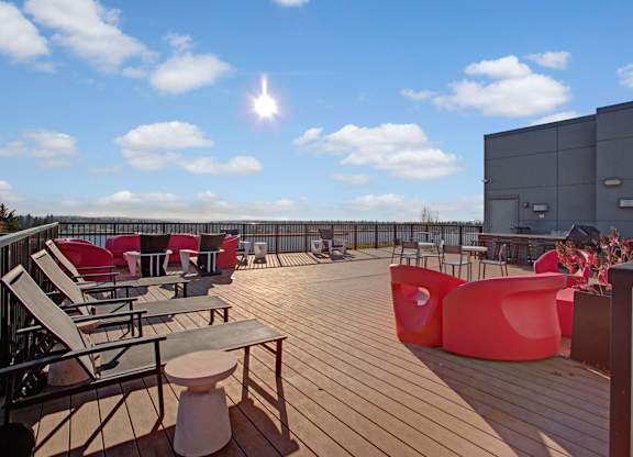 A sunny rooftop deck here at Leilani on Greenwood featuring expansive wood plank flooring, modern lounge chairs arranged for relaxation, bold red sculptural seating, small accent tables, and perimeter railings that frame open sky and distant views, creating an inviting outdoor gathering space designed for socializing and enjoying fresh air.