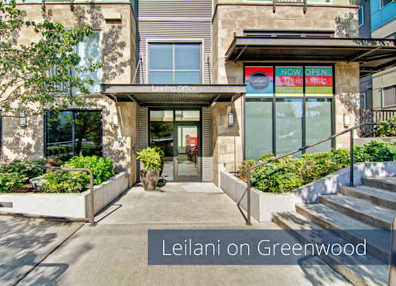 A modern leasing office exterior here at Leilani on Greenwood featuring a glass entry door beneath a metal awning labeled leasing office, large storefront windows with community signage, concrete walkways with handrails, and raised planters filled with greenery that frame the entrance and create a welcoming street-level arrival.