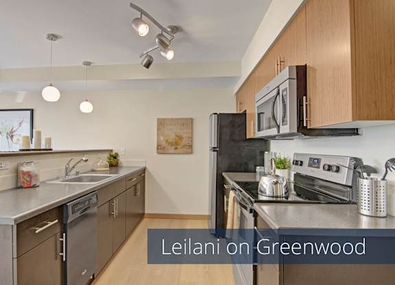 A modern galley-style kitchen here at Leilani on Greenwood featuring flat-panel wood cabinetry, smooth countertops, and a stainless steel sink with a dishwasher below. Dark appliances line the cooking area, while pendant and track lighting, light wood-style flooring, and a breakfast bar create a bright and functional space.