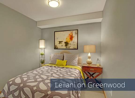 A cozy bedroom here at Leilani on Greenwood featuring soft carpeted flooring, neutral walls, and a bed with patterned bedding and accent pillows. A nightstand with a table lamp, framed artwork above the headboard, and overhead lighting create a warm, comfortable space designed for rest and relaxation.