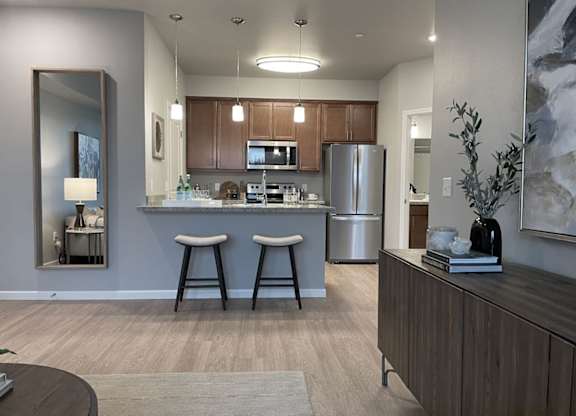 A modern open kitchen and living area here at LIVEasy Street featuring warm wood cabinetry, a stainless-steel refrigerator and range, a breakfast bar with two upholstered stools, pendant lighting, wood-style flooring, and a neutral living space with accent décor and soft natural light creating a welcoming apartment layout.