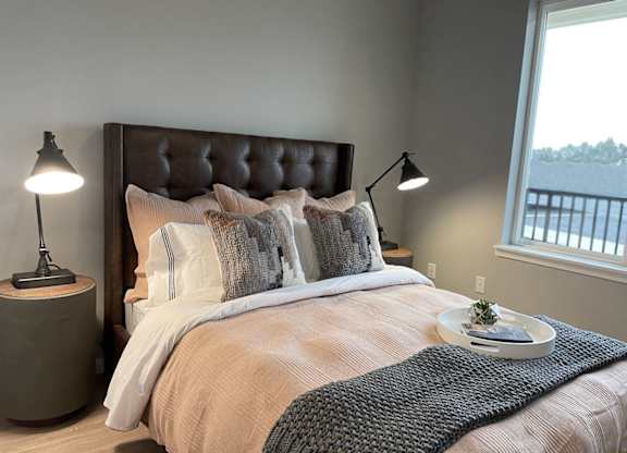 A cozy bedroom here at LIVEasy Street featuring a tufted upholstered headboard, neatly made bed with layered neutral bedding and textured throw, matching bedside tables with modern lamps, light wood-style flooring, and a large window providing natural light and neighborhood views that create a calm, inviting apartment retreat.