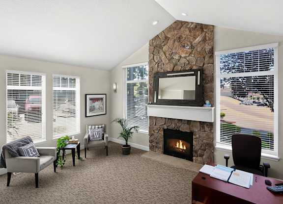 A bright business center here at Regency Woods featuring a stone fireplace with a mantel mirror, large windows with blinds allowing abundant natural light, neutral carpeting, a wooden desk with computer workstation, and cozy accent seating with side tables and plants, creating a comfortable and professional space for work or quiet meetings.