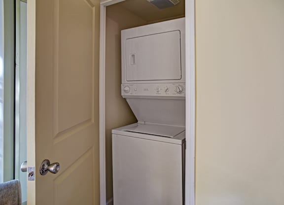 A compact in-home laundry closet here at Regency Woods featuring a stacked washer and dryer neatly tucked behind a paneled door, with neutral wall tones and a space-efficient layout that provides convenient, discreet laundry access within the apartment.