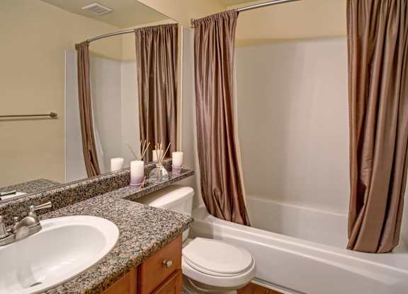 A clean bathroom here at Regency Woods featuring a wide granite-style vanity with an undermount sink, a large wall mirror, wood cabinetry, a toilet, and a tub with a shower curtain, complemented by neutral walls, simple décor accents, and durable flooring for a polished, functional space.