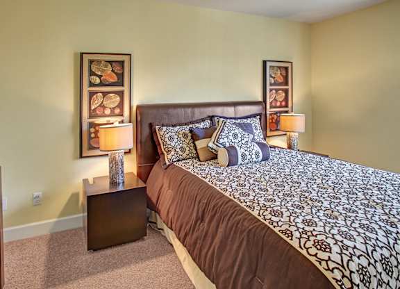 A comfortable bedroom here at Regency Woods featuring a spacious bed with patterned bedding, a padded headboard, matching nightstands with table lamps, framed wall art on either side of the bed, soft carpeted flooring, and light neutral walls that create a calm and inviting atmosphere.