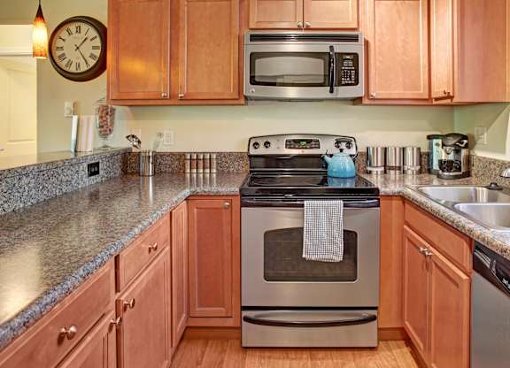 A warm, inviting kitchen here at Regency Woods featuring wood cabinetry, speckled countertops, and a stainless-style oven with a built-in microwave above, complemented by a double-basin sink, dishwasher, small appliances on the counters, and wood-style flooring that creates a functional and welcoming cooking space.