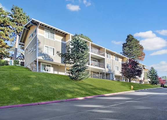 A hillside apartment building here at Regency Woods featuring multi-level residences with private balconies, neutral-toned siding, and large windows, set above a gently sloped green lawn with mature trees, seasonal foliage, and a paved driveway below under a clear blue sky with soft clouds.