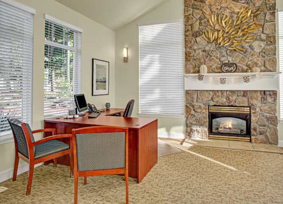 A welcoming business center here at Regency Woods featuring a wooden desk with seating, computer workstation, and large windows with blinds, paired with a stone fireplace and decorative mantel, neutral carpeting, and soft natural light that creates a comfortable, professional space for work or study.