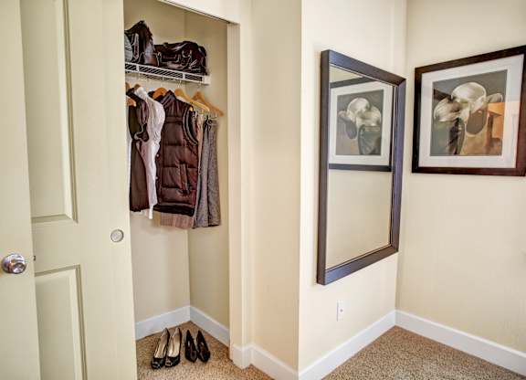 A compact entry closet area here at Regency Woods featuring an open hanging rod with shelving above, neatly arranged coats and jackets, carpeted flooring, and a recessed nook beside a full-length wall mirror and framed artwork, offering convenient storage and a clean, organized transition space near the entry.