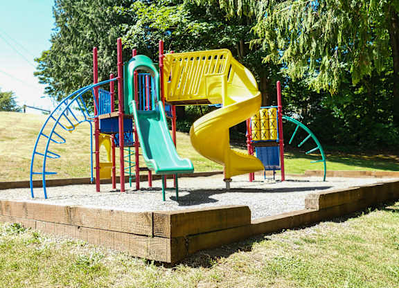 A colorful outdoor playground here at Regency Woods featuring a raised play structure with red support posts, a green straight slide, a yellow spiral slide, climbing ladders, and activity panels, all set on a gravel play surface bordered by timber edging and surrounded by open grassy areas and mature evergreen trees.