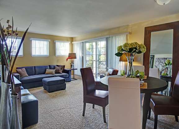 A spacious living and dining area here at Regency Woods featuring plush carpet flooring, a sectional sofa with an ottoman, a round dining table with mixed upholstered chairs, large windows with vertical blinds, and a sliding glass door leading to a private balcony, creating a warm, open layout filled with natural light.