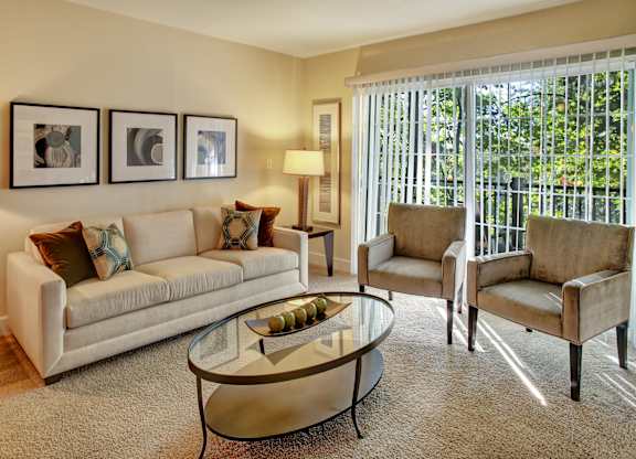 A bright living room here at Regency Woods featuring a neutral sofa with patterned accent pillows, two upholstered armchairs, and a glass coffee table set on plush carpeting, with framed wall art, a floor lamp, and large sliding glass doors with vertical blinds that open to a private balcony and bring in natural light.