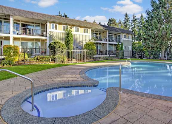 A serene outdoor swimming pool here at Regency Woods featuring a curved pool design with a shallow entry area, metal handrails, and smooth concrete decking, set against landscaped greenery, manicured grass, mature trees, and multi-story apartment buildings with balconies under a bright blue sky.