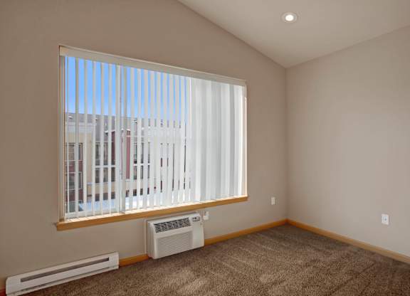 A bright bedroom here at Riverside 9 with soft carpet, large window with vertical blinds, and built-in air conditioning for comfort and natural light.